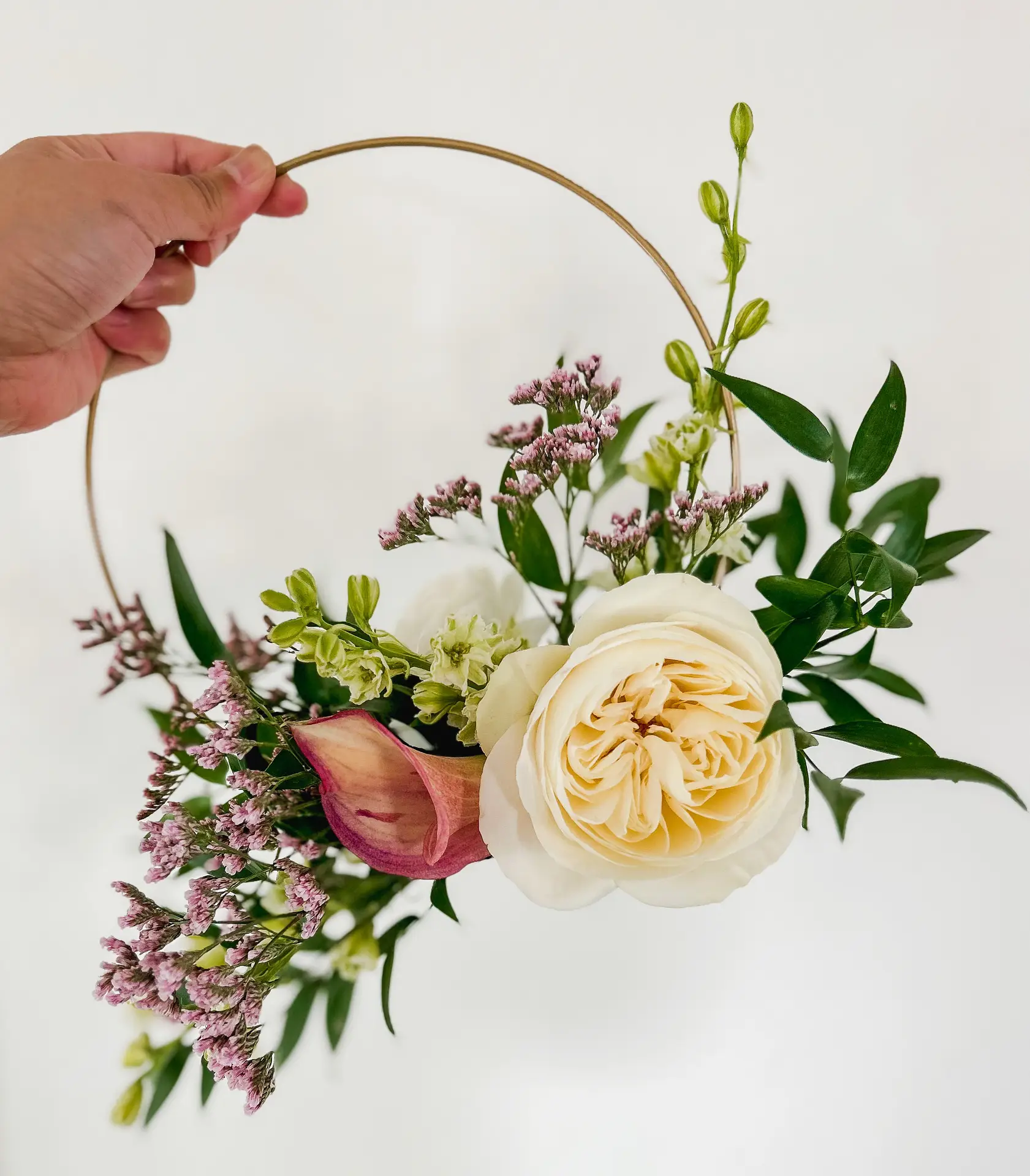 Hand holding a floral hoop with a cream garden rose, burgundy calla lily, pink limonium and greenery