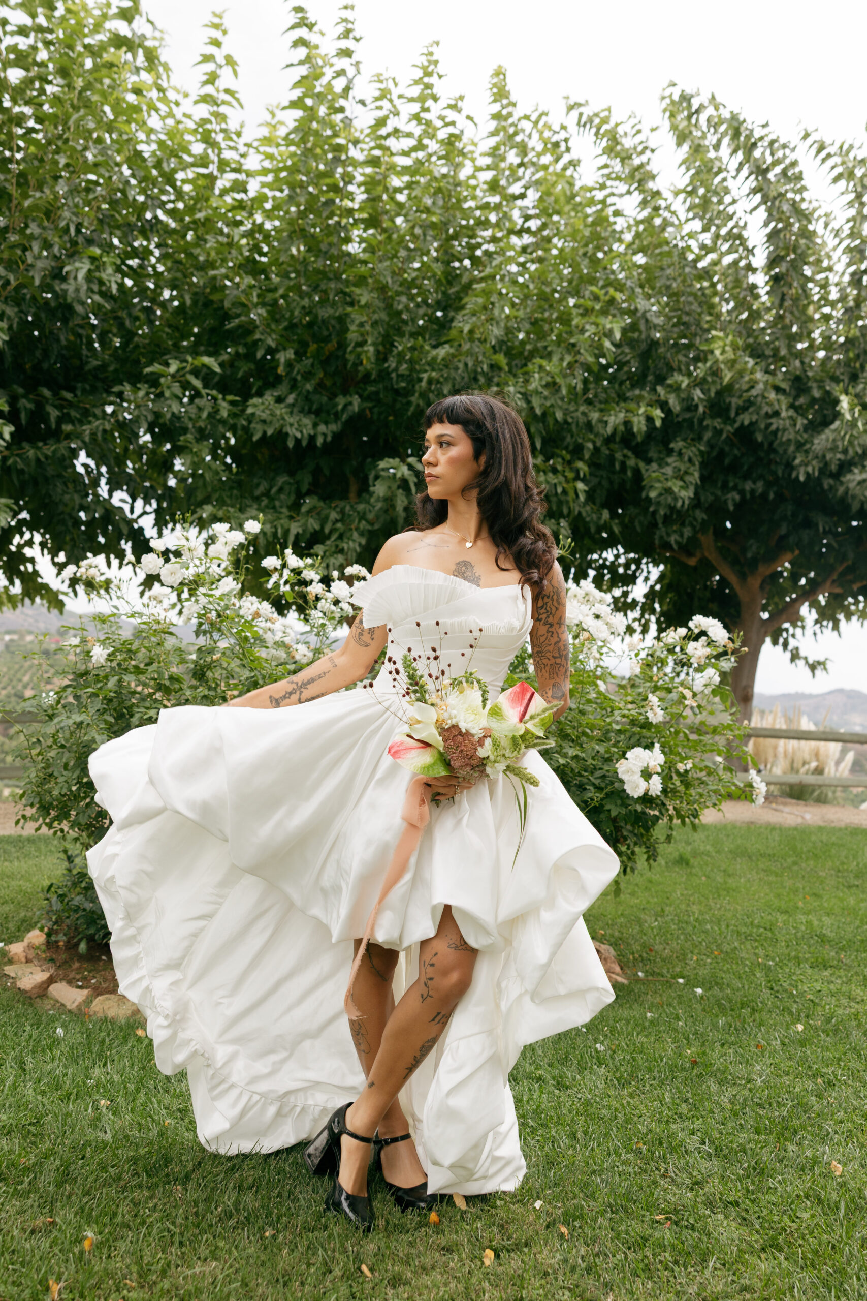 Bride in a sculptural gown holding a calla lily and anthurium bouquet near garden roses