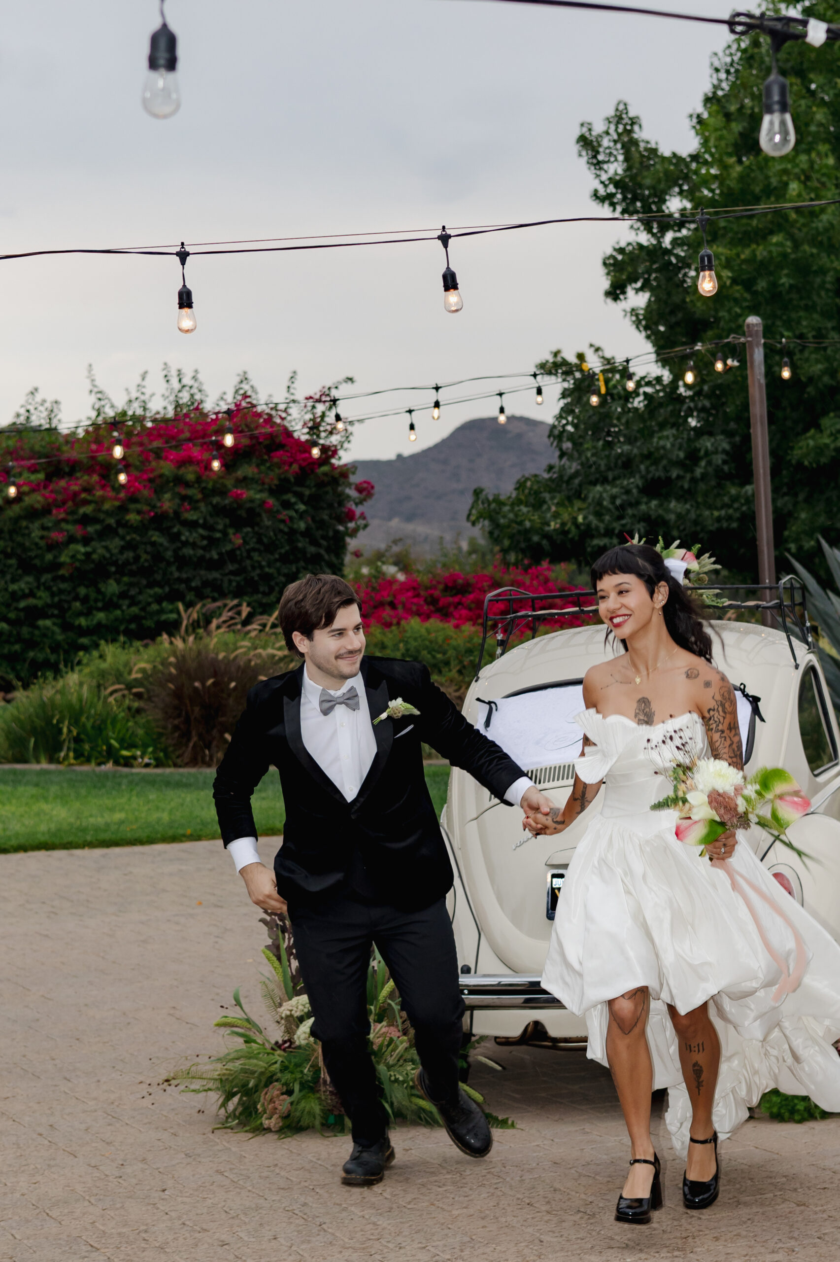 Newlyweds walking hand in hand past a vintage car strung with string lights