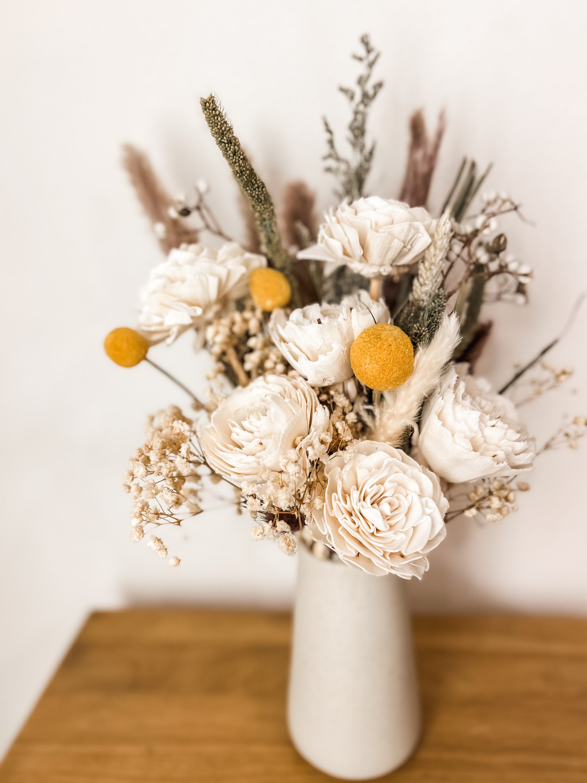 Neutral dried-flower arrangement with cream roses, yellow craspedia, and dried grasses in a white ceramic vase