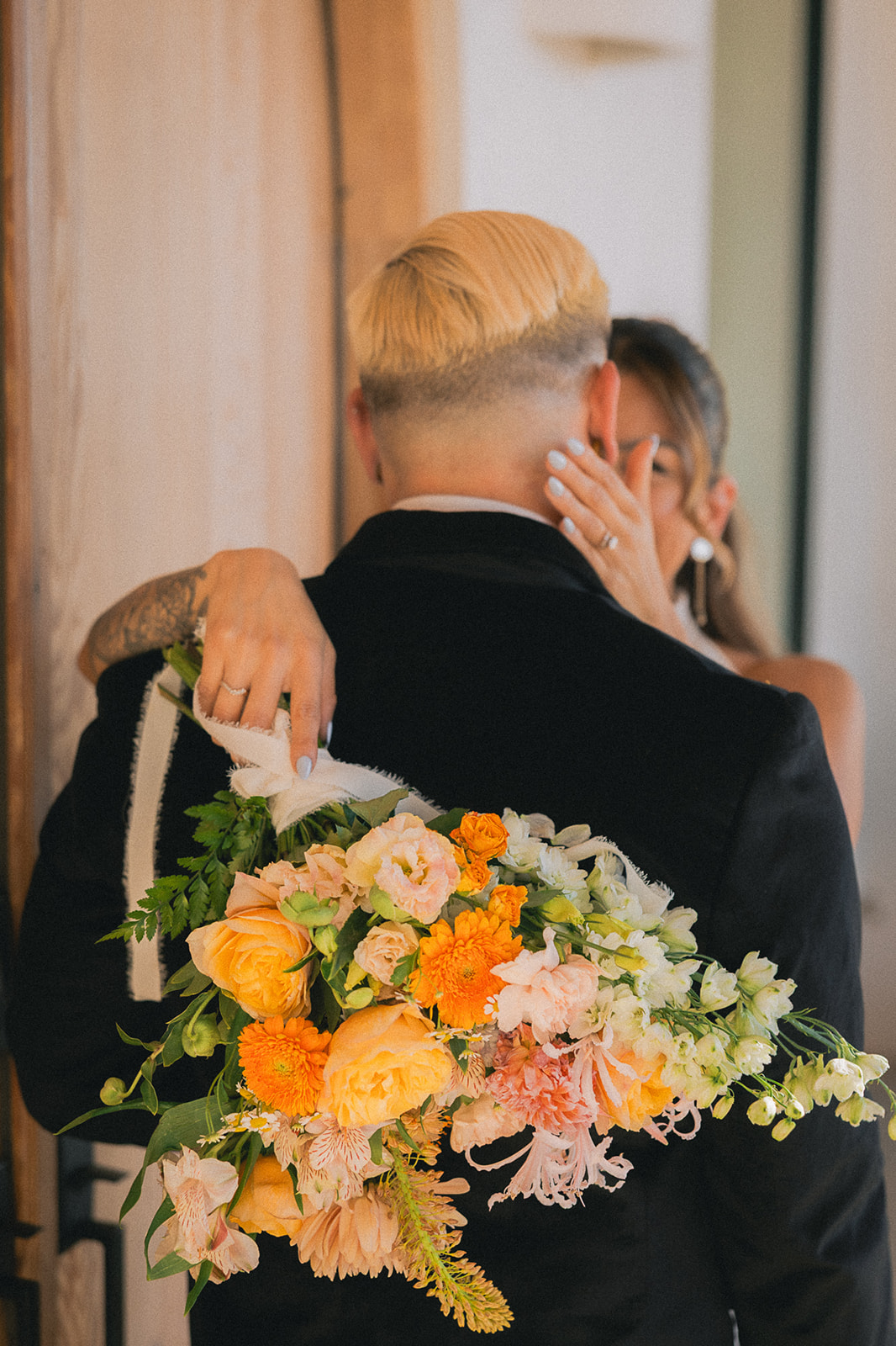 Groom embraces his bride while holding her peach and orange garden bouquet, designed through full-service wedding florist services