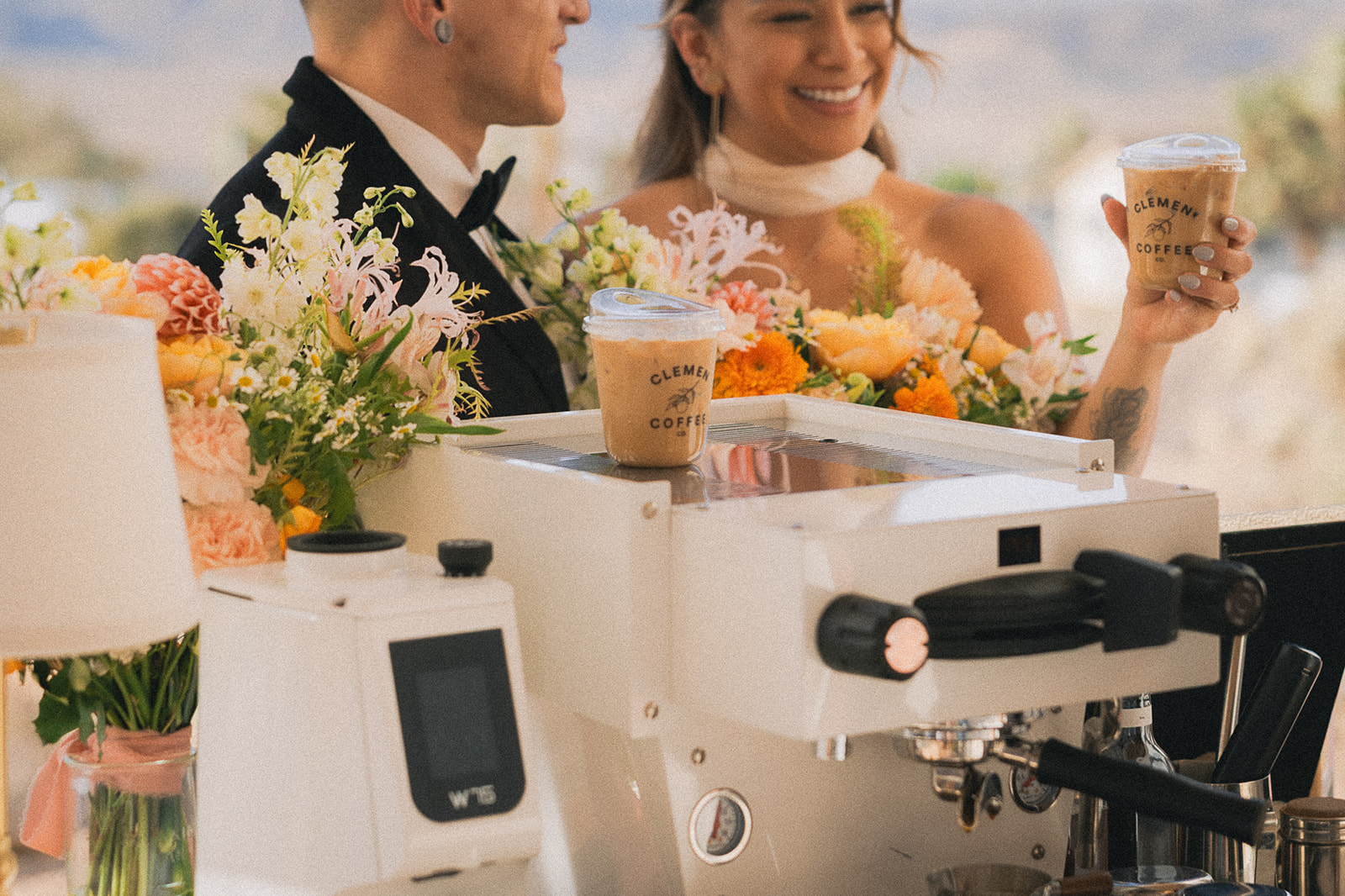 Bride and groom hold iced coffees at an espresso cart decorated with blooms, a reception detail covered by wedding florist services