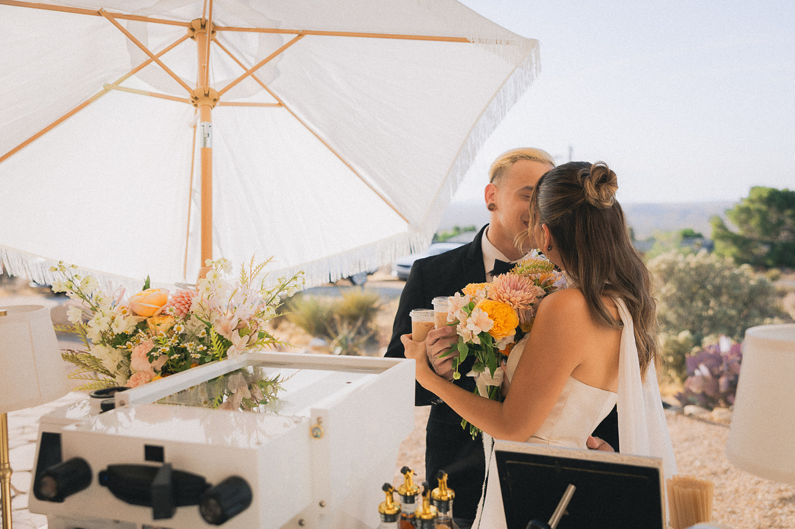 Bride and groom embrace beside a floral-trimmed espresso cart under a white umbrella with desert views