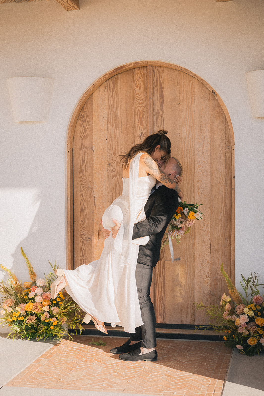 Groom lifts his bride in front of an arched wooden door framed by ground floral arrangements from wedding florist services