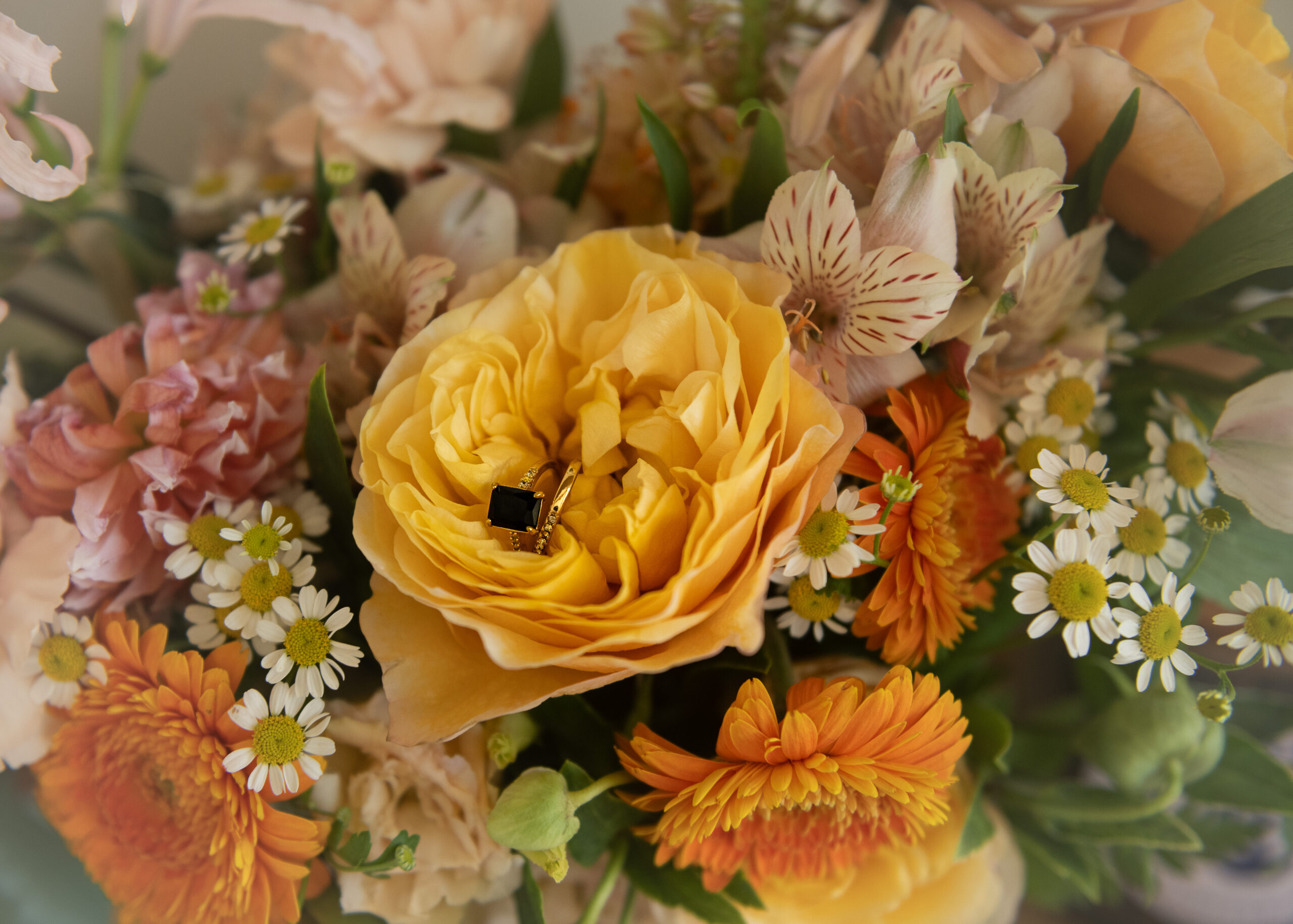Wedding ring set on a golden garden rose, with chamomile daisies, alstroemeria, and orange blooms around it