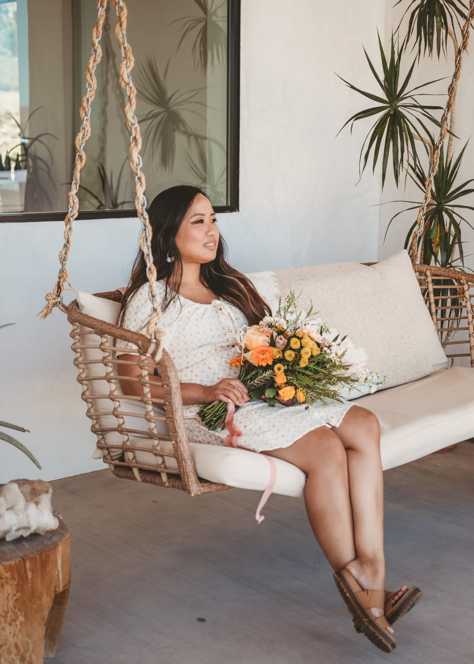 Woman in a white dress smiles on a rattan swing holding a bouquet from Sage & Autumn's wedding florist services