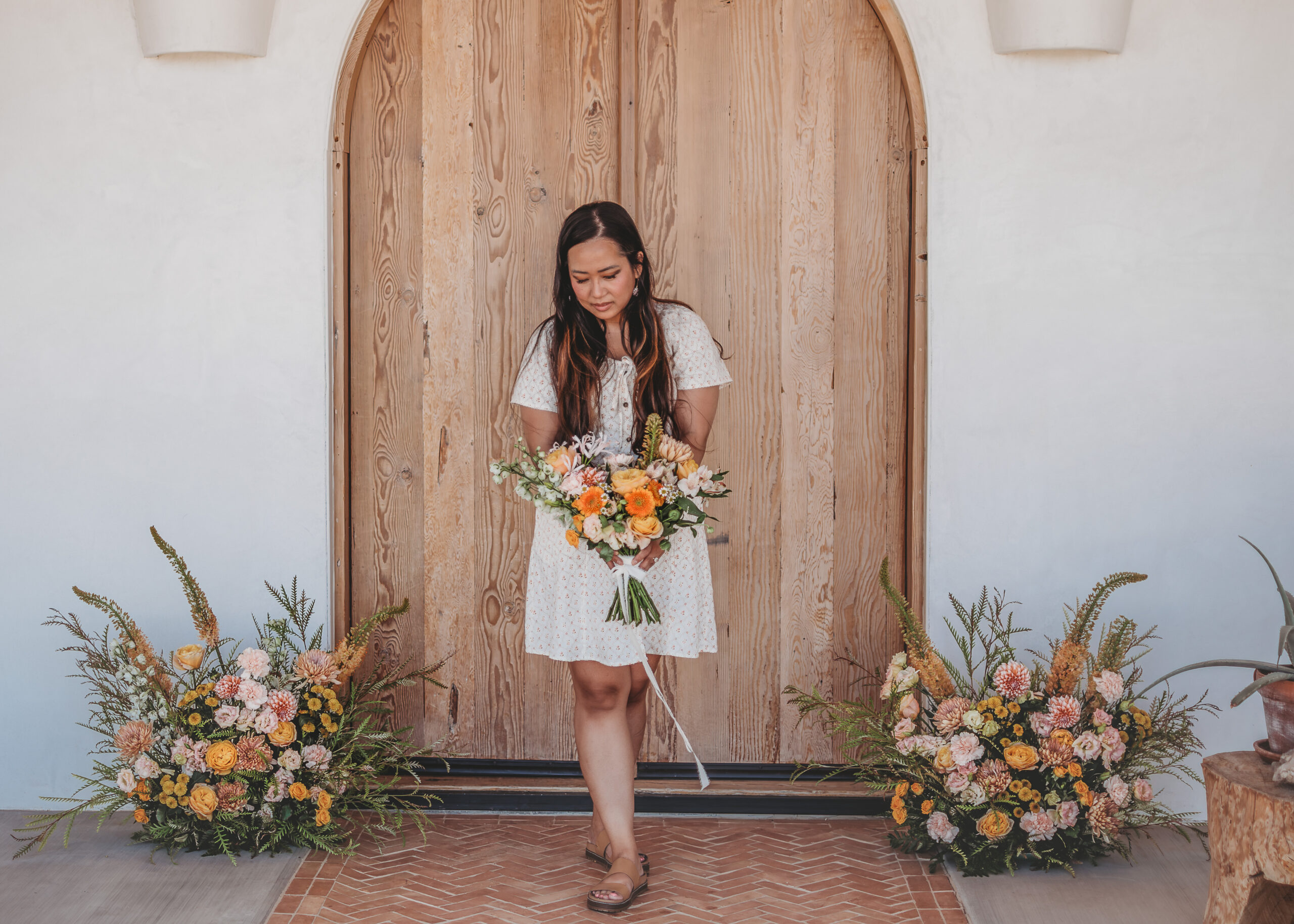 Woman in a white dress holds a garden bouquet between two meadow-style ground arrangements, an example of wedding florist services