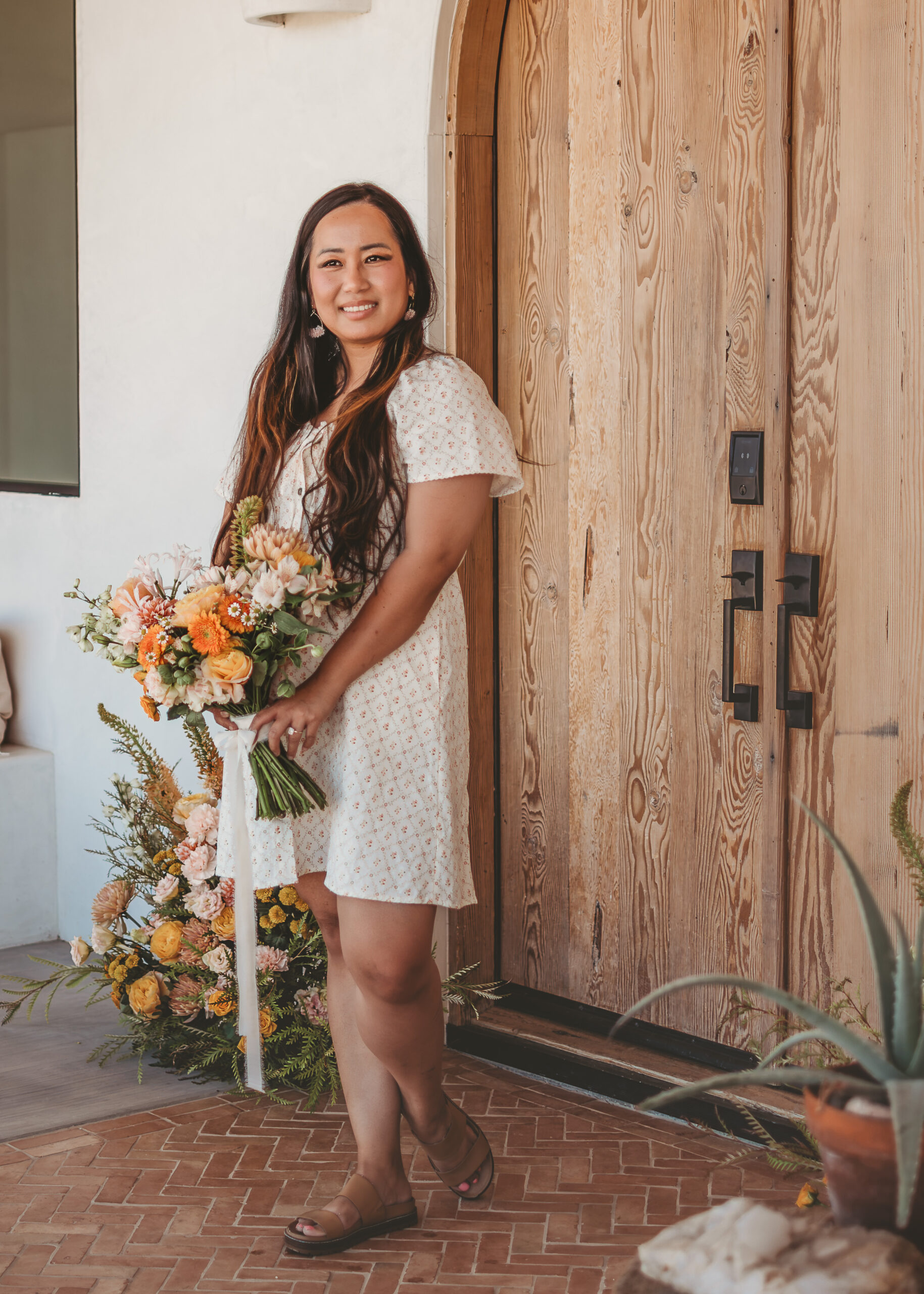 Smiling woman in a white eyelet dress holds a peach, orange, and blush bouquet with trailing ribbon beside an arched wooden door
