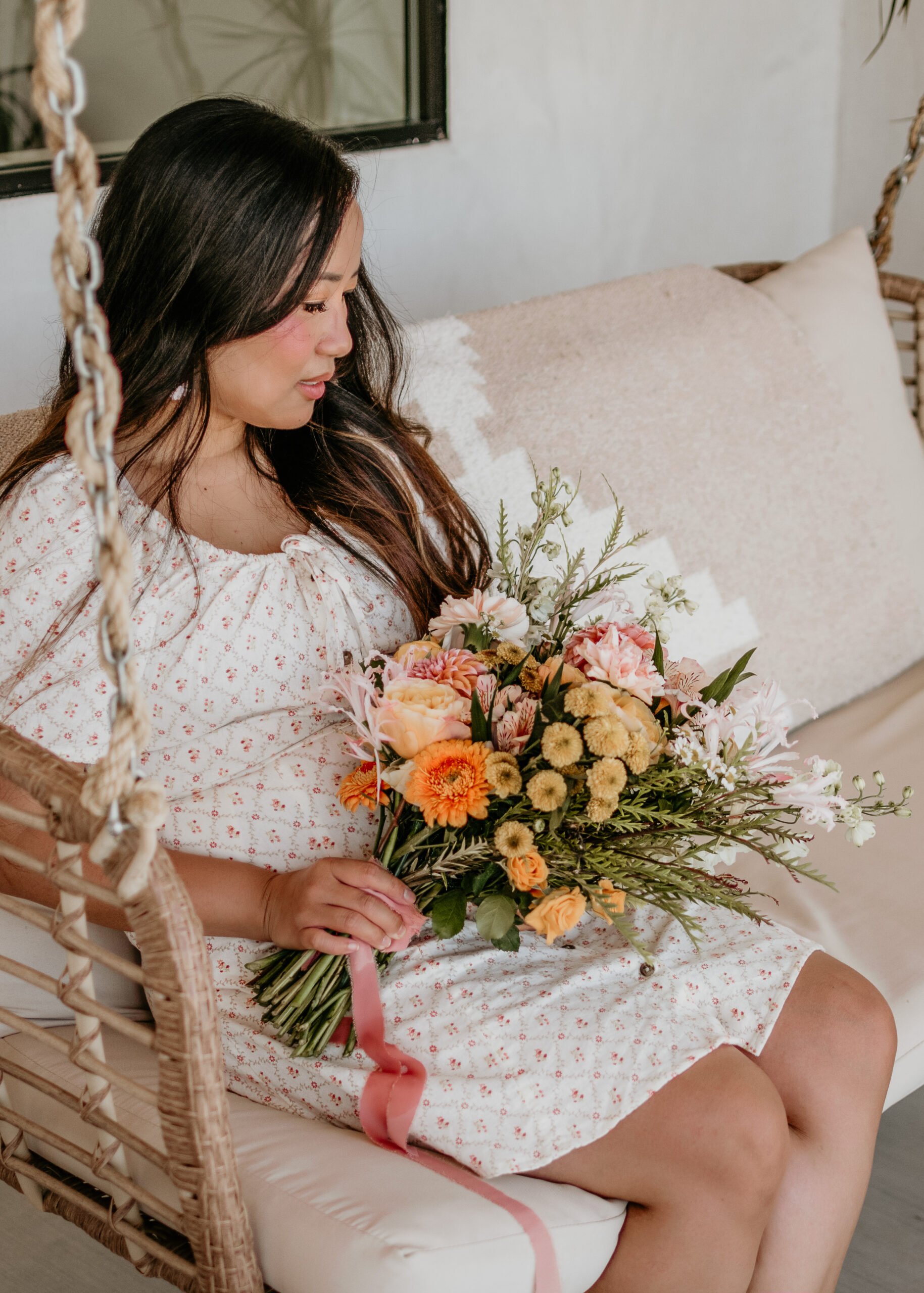 Woman in a floral dress seated on a rattan hanging swing, holding a garden bouquet of orange, peach, and blush flowers